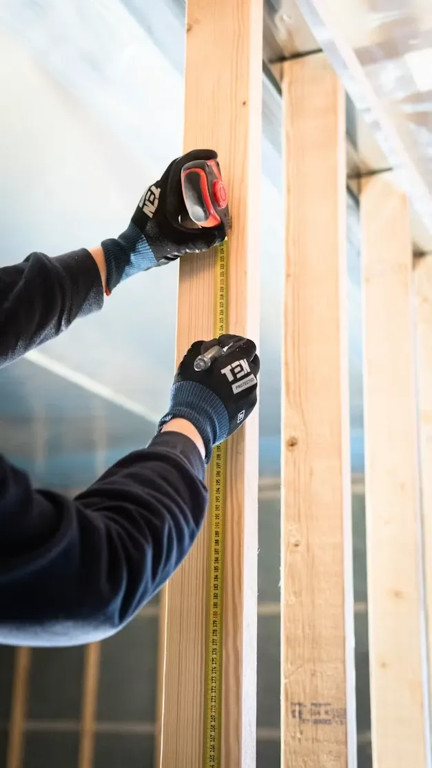 Tømrer med arbeidshansker måler en stender med målebånd